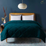 A teal quilted comforter on a bed in a room with a blue wall and a hanging lamp, alongside a wooden nightstand with a plant and a book.